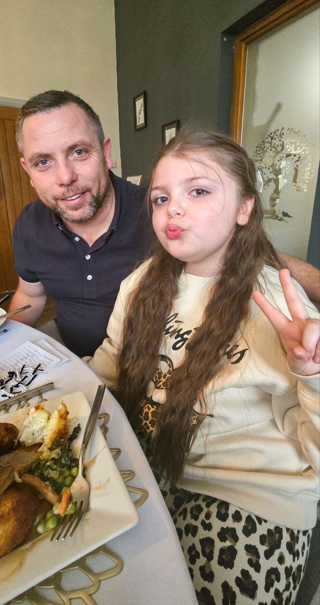 Stephen and Daisy seated at dining table with roast dinner, Daisy posing with peace sign
