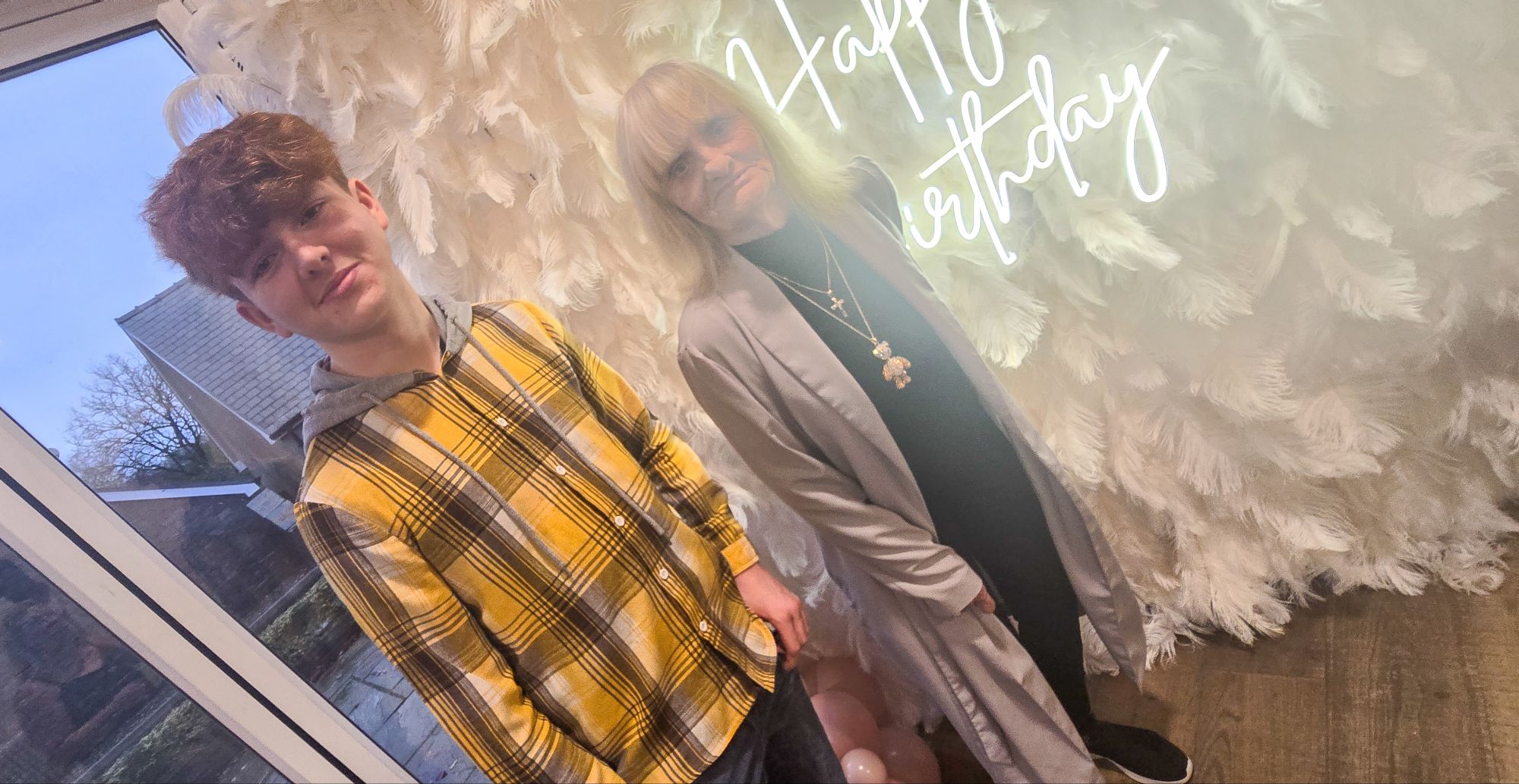 Mum and grandson Zack in front of feather wall with birthday neon sign