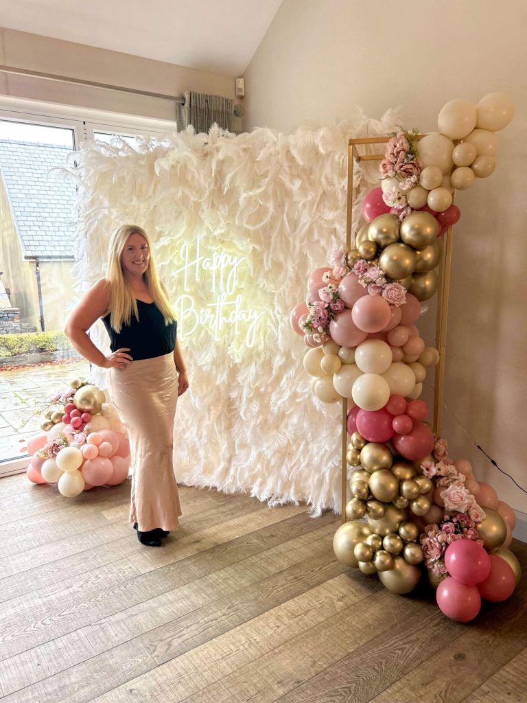 Corrina posing in front of feather wall with birthday balloons and neon sign