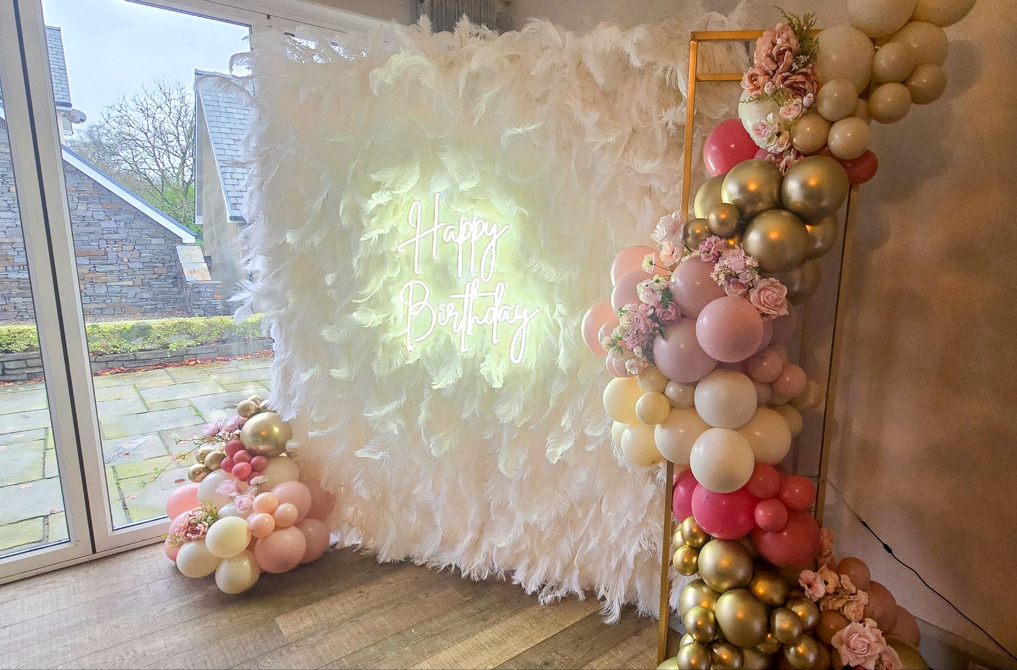 Feather wall with glowing “Happy Birthday” neon sign and floral balloon garland