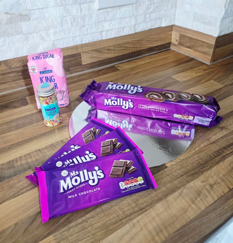 Before shot of Yule log ingredients on a kitchen worktop, including a silver cake board, Molly's chocolate Swiss roll in purple packaging, three packets of Molly's milk chocolate, a bag of icing sugar, and a jar of celebration sprinkles.