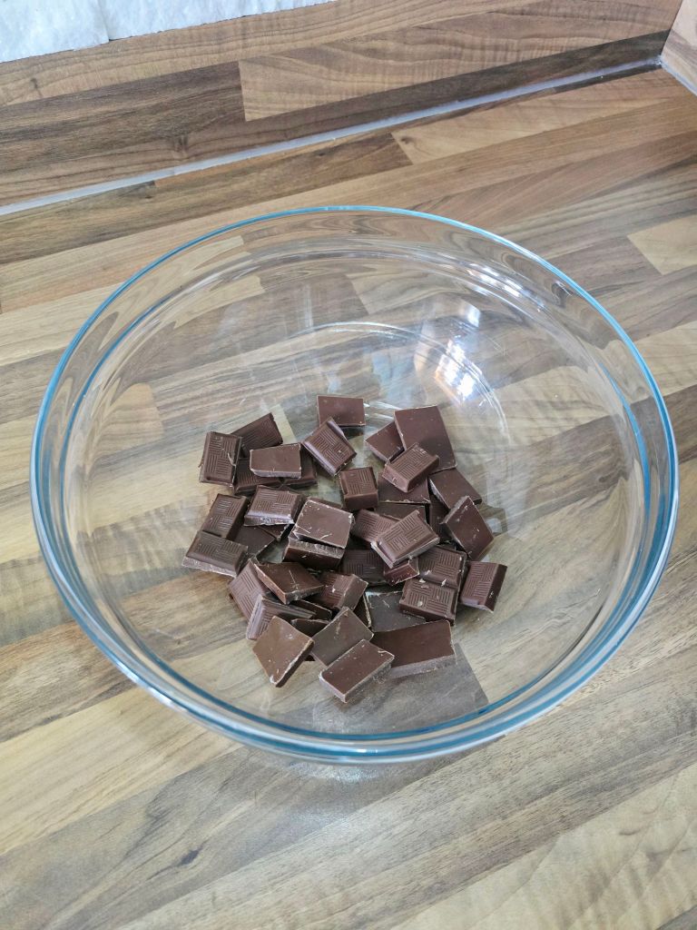 Top down view of a large glass bowl on a kitchen worktop filled with Molly's milk chocolate broken into small square pieces, ready for melting.