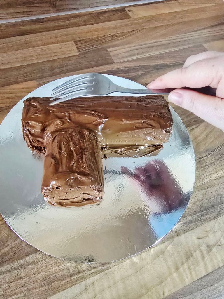 Chocolate covered Yule log on a kitchen worktop with a shiny, tempered finish, and a hand holding a fork ready to create wood grain lines.