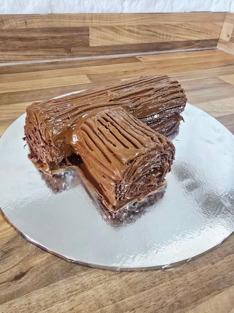 Yule log coated in glossy chocolate with fork created wood grain lines, sitting on a silver cake board on a kitchen worktop, ready for decorating.