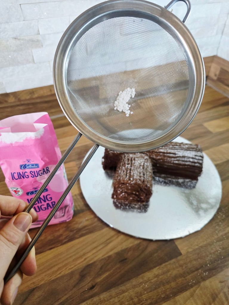 Large stainless steel sieve with icing sugar inside, placed on a kitchen worktop. Out of focus underneath is a chocolate covered Yule log already dusted with icing sugar for a snowy effect, with a bag of icing sugar to the left.