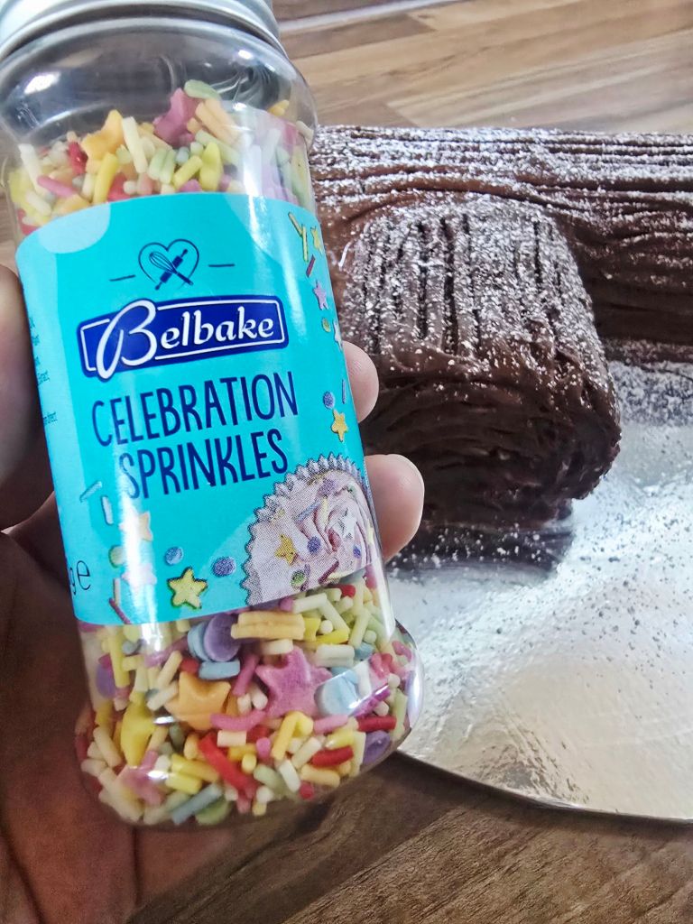Close up of a hand holding a Belbake Celebration Sprinkles jar showing colourful sprinkles inside, with a snowy chocolate Yule log slightly blurred in the background on the kitchen worktop.