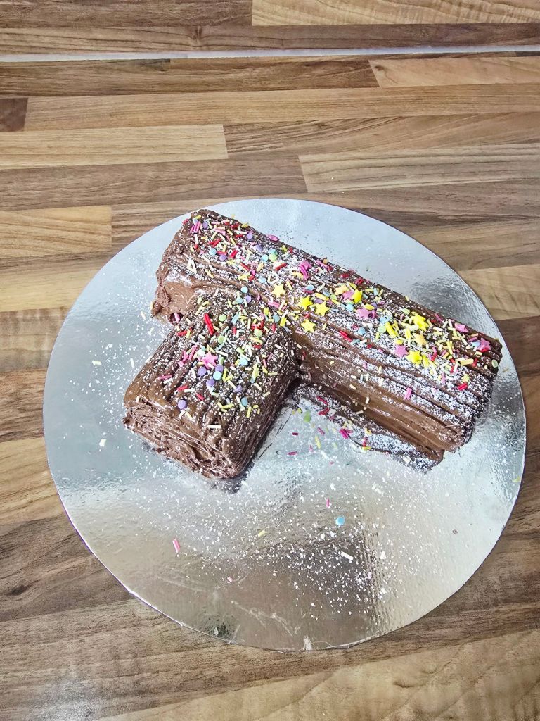 Aerial view of a chocolate covered Yule log on a silver cake board, dusted with icing sugar for a snowy effect and topped with colourful sprinkles, sitting on a kitchen worktop.