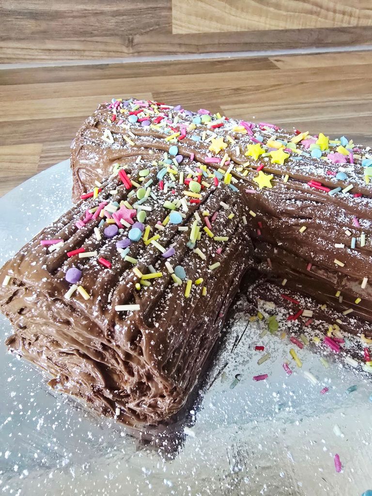 Close up view of a chocolate covered Yule log showing the branch piece sticking out, decorated with colourful sprinkles and part of the main log visible on a silver cake board.