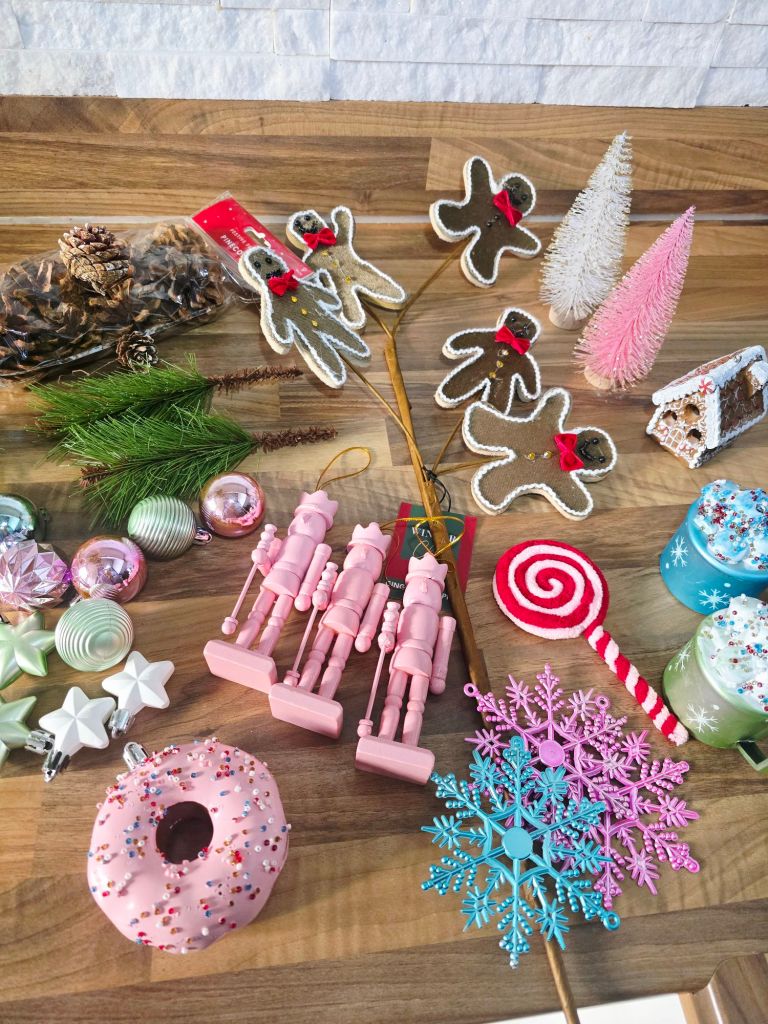 Top down view of assorted Yule log decorations on a kitchen worktop, including a packet of pine cones, artificial pine needles, five gingerbread men picks, two pastel bottle brush trees, a miniature gingerbread house, two mugs of hot cocoa decorations (blue and green) with whipped cream and colourful toppings, a red and pink striped lollipop, three pink nutcrackers, blue and pink plastic snowflakes, a pink plastic doughnut with sprinkles, and assorted baubles and star shaped ornaments in pink, green, blue, and white.