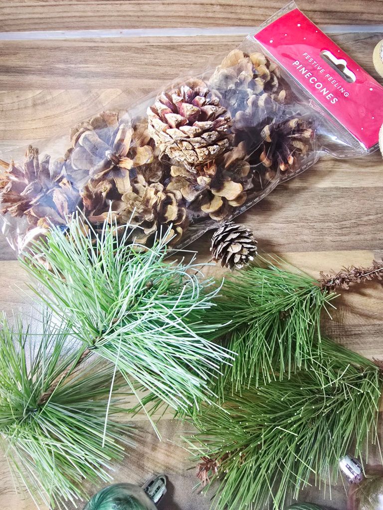 Close up of a sealed packet of pine cones alongside artificial green Christmas tree pine needles on a kitchen worktop.