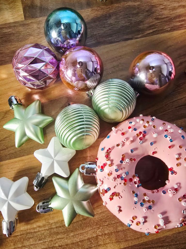 Top down view of assorted Yule log decorations on a kitchen worktop, including a pink doughnut, two white star shaped baubles, two green star shaped baubles, two green mini baubles, and four textured baubles in pink and blue.