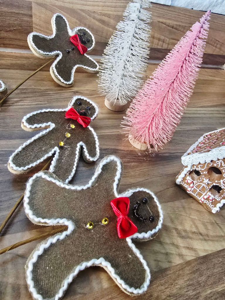Close up of Yule log decorations on a kitchen worktop, including three gingerbread men on picks, white and pink pastel bottle brush trees, and a miniature gingerbread house partially visible.