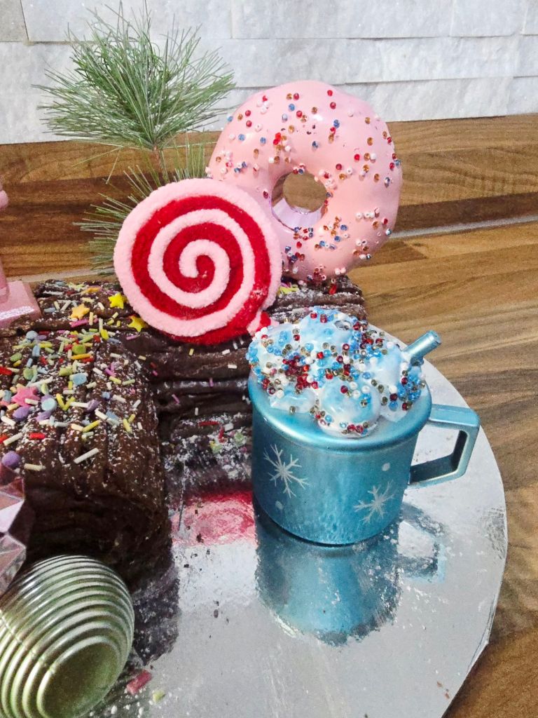 Close up of a chocolate Yule log decorated with a blue hot cocoa mug, a red and pink striped lollipop, and a pink doughnut with sprinkles, with a green pine needle and a mini green bauble visible on the side.