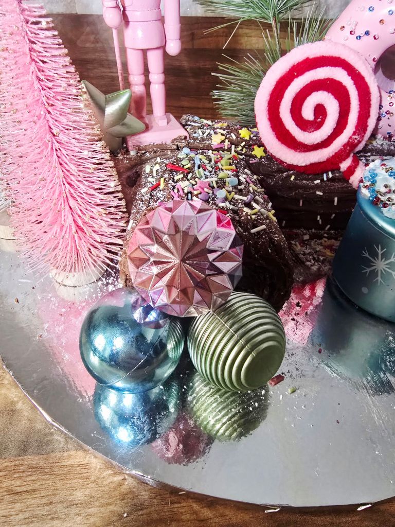 Close up of three mini baubles in blue, pink, and green placed on a chocolate Yule log, with the bottom half of a pink bottle brush tree, the base of a pink nutcracker, the head of a striped lollipop, and a hint of green pine needle visible.