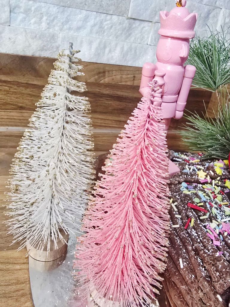 Close up view of two pastel bottle brush trees in pink and white next to a chocolate Yule log, with a pink nutcracker sitting on top in the background, sprinkles on the log, and a hint of green pine needles visible.