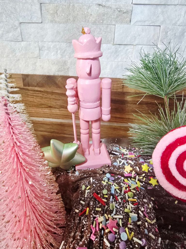 Close up of a chocolate Yule log with a pink nutcracker standing on top, green pine needle to the right, the top of a pink and red striped lollipop visible, sprinkles on the log, and a mini star shaped bauble with a pink bottle brush tree at the nutcracker’s base.