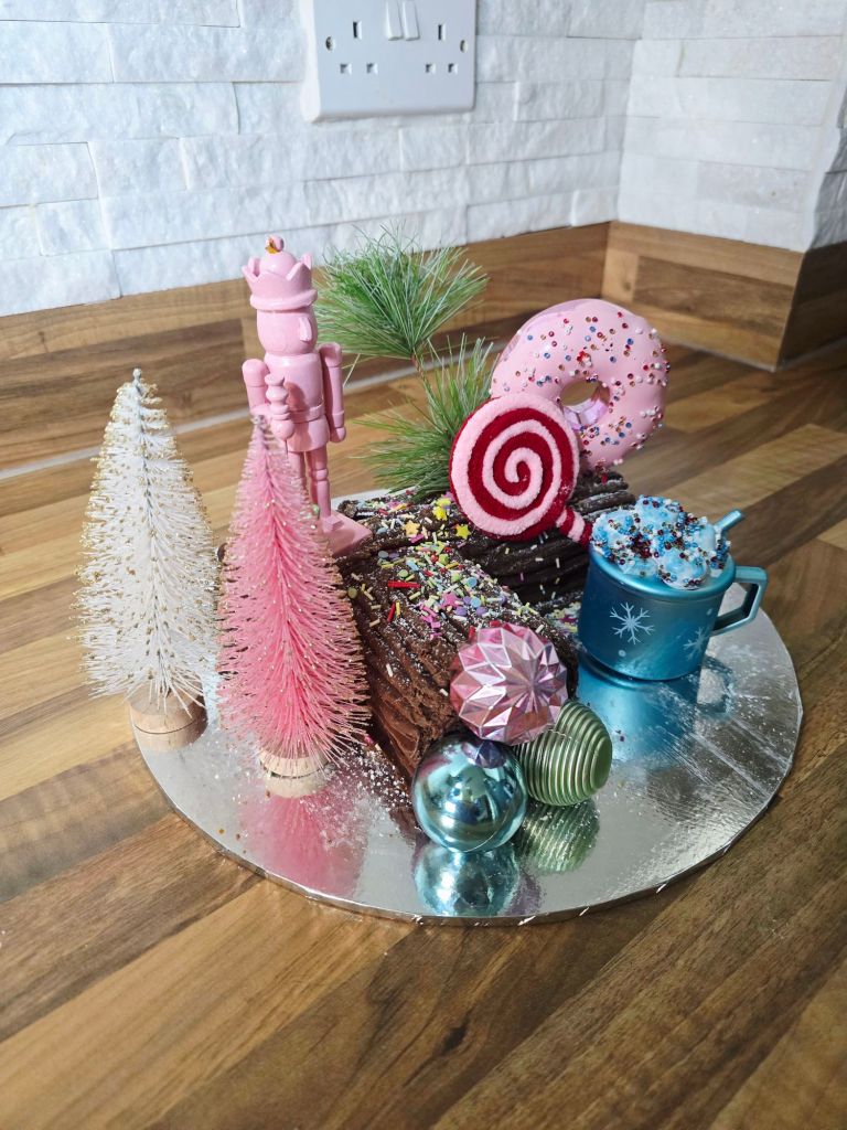 Chocolate Yule log covered in glossy chocolate and sprinkles, decorated with pastel bottle brush trees, pink nutcracker, pink doughnut, blue cocoa mug, striped lollipop, and colourful baubles.