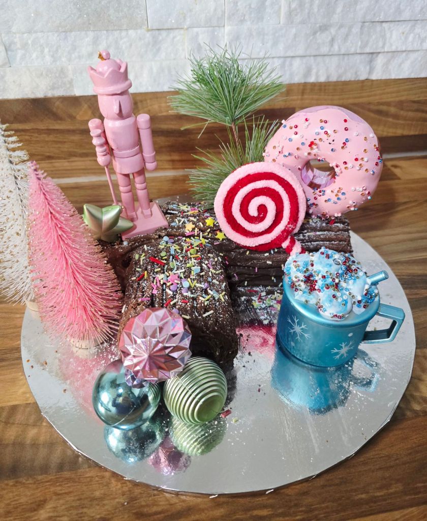 Overhead view of a Candy Land inspired Yule log featuring pink and white trees, pink nutcracker, doughnut with sprinkles, blue cocoa mug, striped lollipop, and assorted pastel baubles.